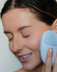 Woman using a blue facial cleansing brush on her face against a neutral background