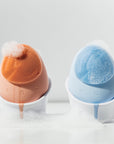 Two cleansing brushes, one orange and one blue, with white bases on a light gray background.