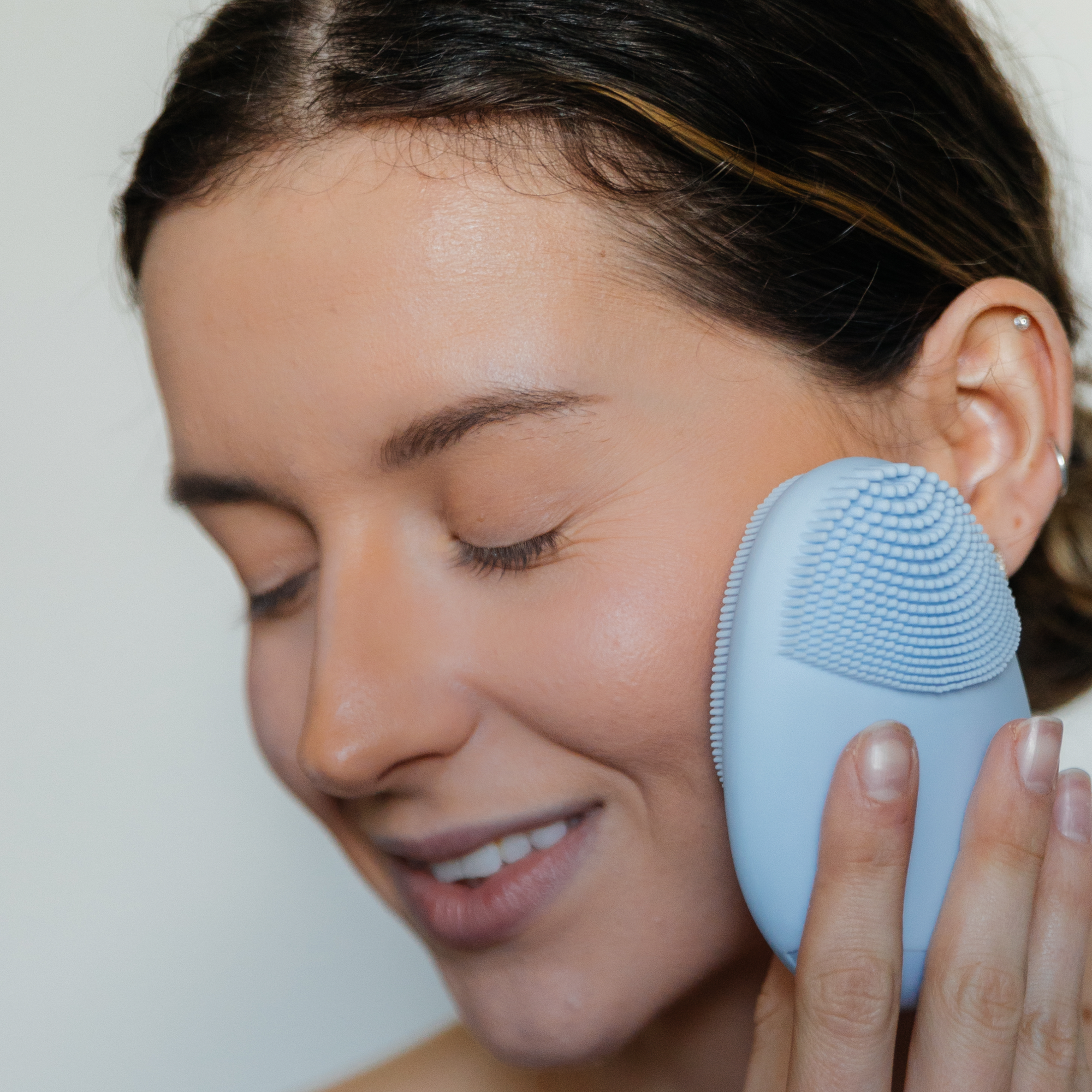 Woman using a blue facial cleansing brush on her face against a neutral background