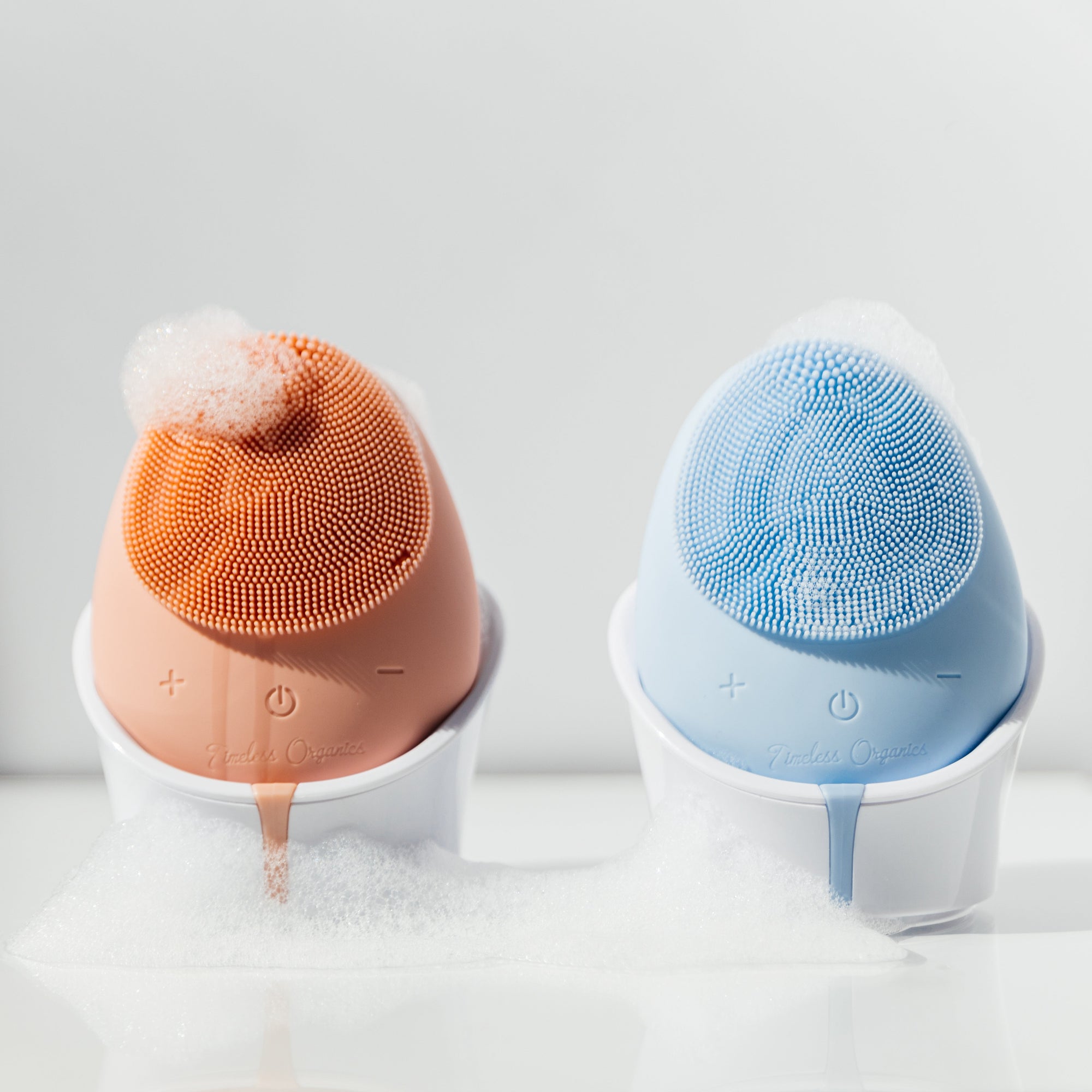 Two cleansing brushes, one orange and one blue, with white bases on a light gray background.